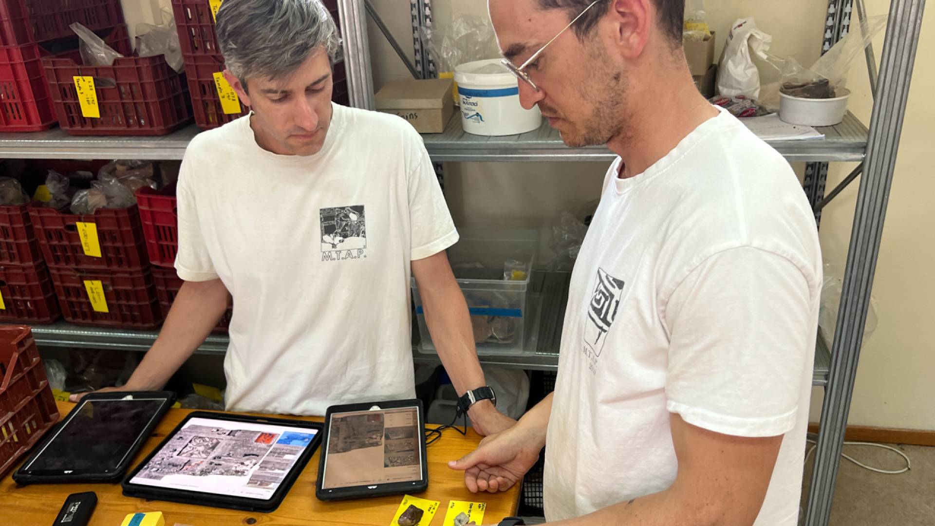 Two professors using electronic devices while examining excavated fragments.